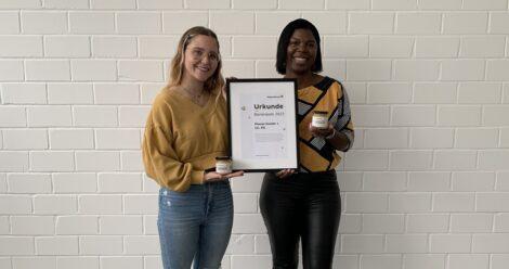 Zwei Planol Mitarbeiter vor weißer Steinwand mit Urkunde zur Bienenpatenschaft und Honig im Rahmen der Ecoplan-Serie von Planol