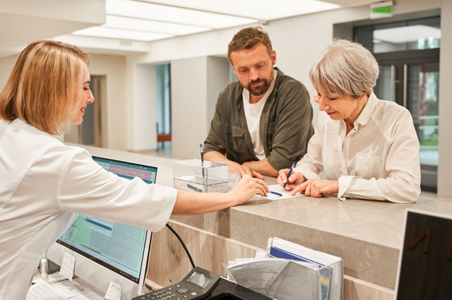 Patientin unterschreibt am Krankenhausempfang, Piepenbrock Mitarbeiterin berät, Begleiter steht daneben.**