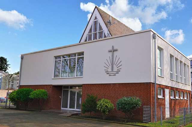 Zweistöckiges Kirchengebäude mit Kreuzsymbol an der Fassade, rotem Klinkersockel und begrüntem Außenbereich