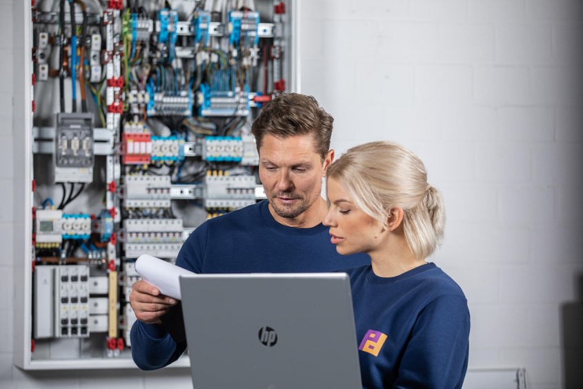 Ein Mann und eine Frau stehen vor einem Schaltkasten und prüfen Dokumente auf einem Laptop