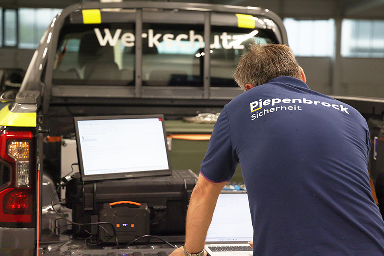 Piepenbrock Mitarbeiter prüft Sicherheitstechnik an Laptops neben einem Werkschutzfahrzeug