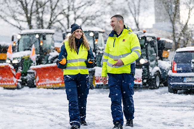 Zwei Piepenbrock Winterdienst-Mitarbeiter in Warnjacken im Schnee vor Räumfahrzeugen