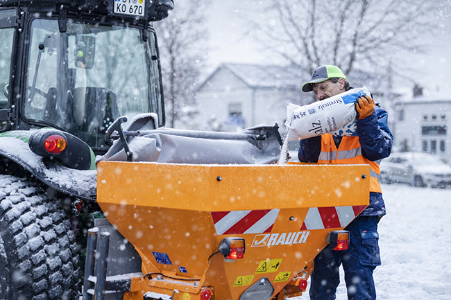 Piepenbrock Mitarbeiter füllt Streusalz in Streugerät am Traktor, während dichter Schneefall die Umgebung bedeckt.