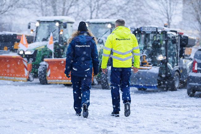 Zwei Piepenbrock Mitarbeiter laufen bei Schneefall an Räumfahrzeugen und Traktoren vorbei.