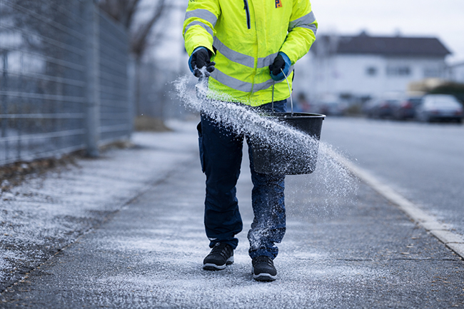 Piepenbrock Mitarbeiter streut mit Eimer Salz auf vereistem Gehweg entlang einer Straße im Winter.