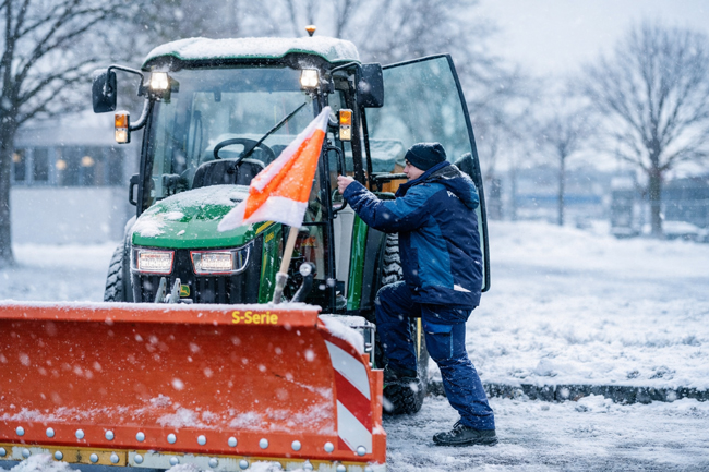 iepenbrock Mitarbeiter steigt bei Schneefall in Traktor mit Schneeschild ein, bereit zum Räumen der Fläche.